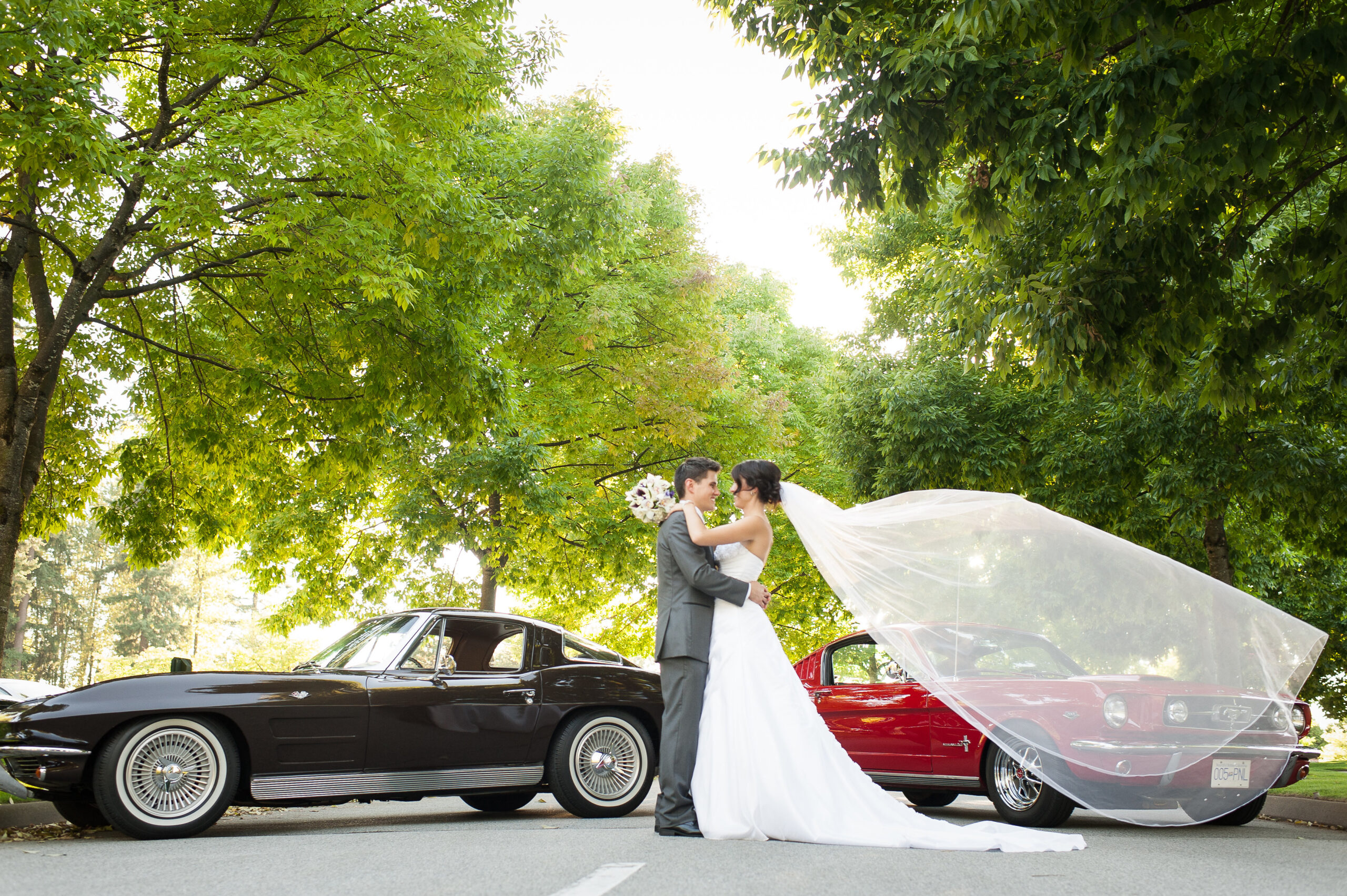 Bride and groom embracing each other with two vintage cars behind them